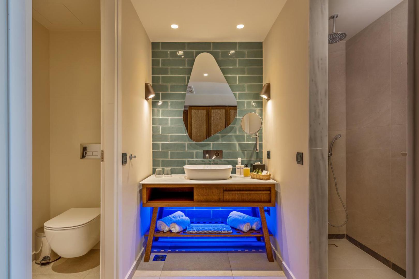 Minos Ambassador Suites & Spa dizaina vannas numurs ar tirkīza flīzēm, asimetrisku spoguli un atsevišķu dušas zonu