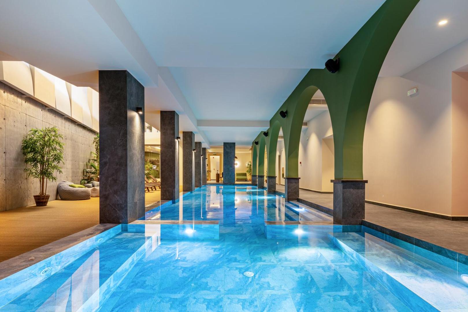 Minos Ambassador Suites & Spa iekštelpu baseins ar arkām, tumšām kolonnām un relaksācijas zonu spa centrā