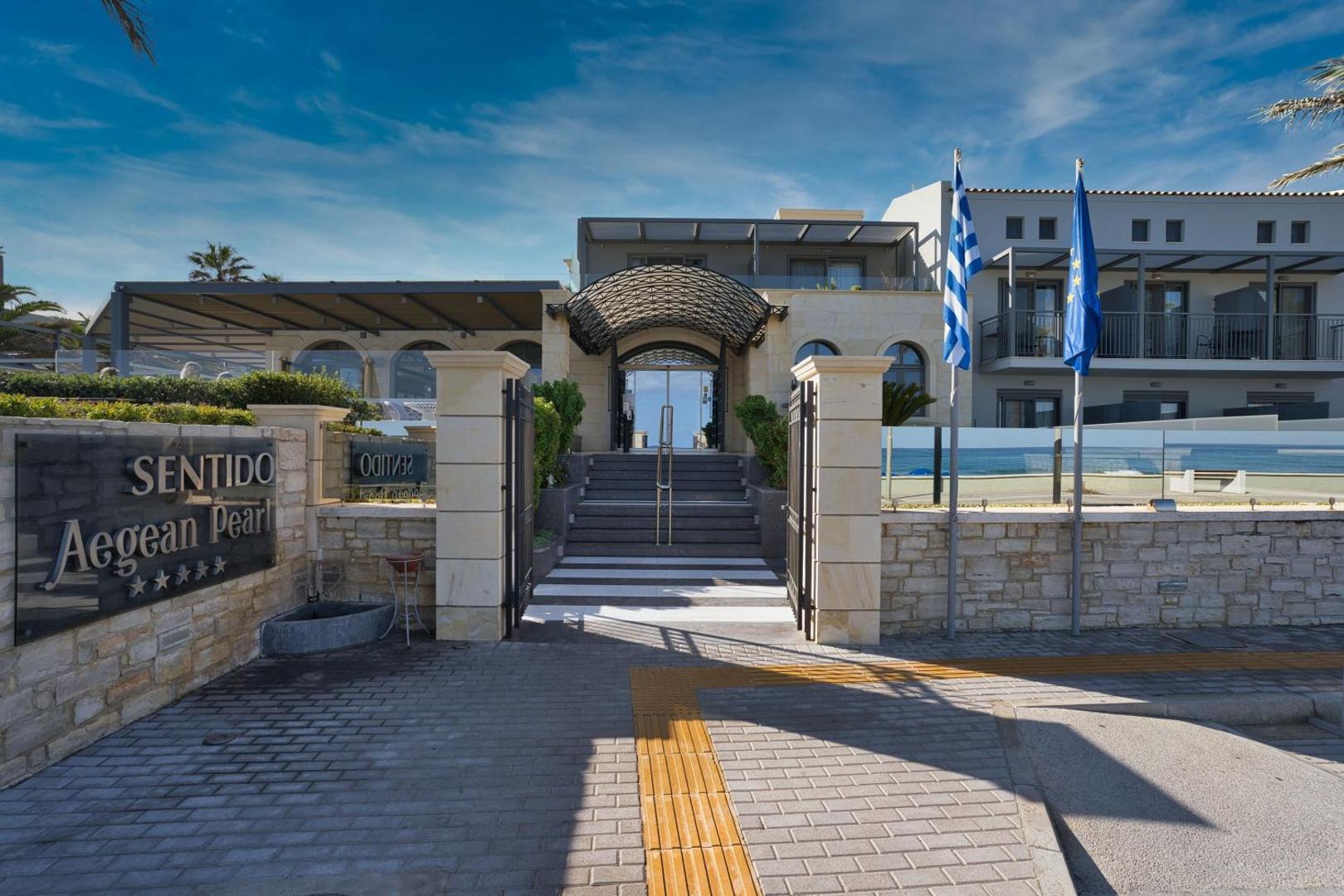Sentido Aegean Pearl galvenā ieeja ar akmens apdari, karogiem un kāpnēm, kas ved uz modernu recepcijas zonu