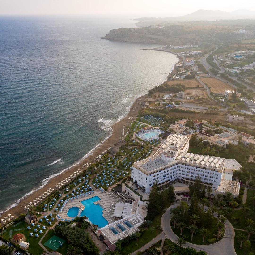 Creta Star Hotel piekrastes ainava no augšas — viesnīcas ēka, zilie baseini un līkumotā Krētas salas krasta līnija saulrietā