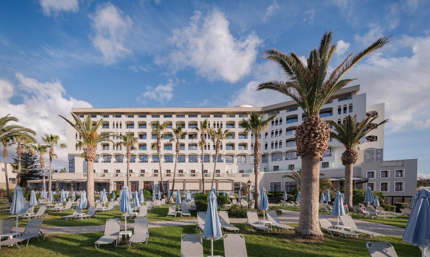 Creta Star Hotel galvenā ēka un dārzs ar palmām — sauļošanās krēsli uz zaļā zālāja un gaiša viesnīcas fasāde
