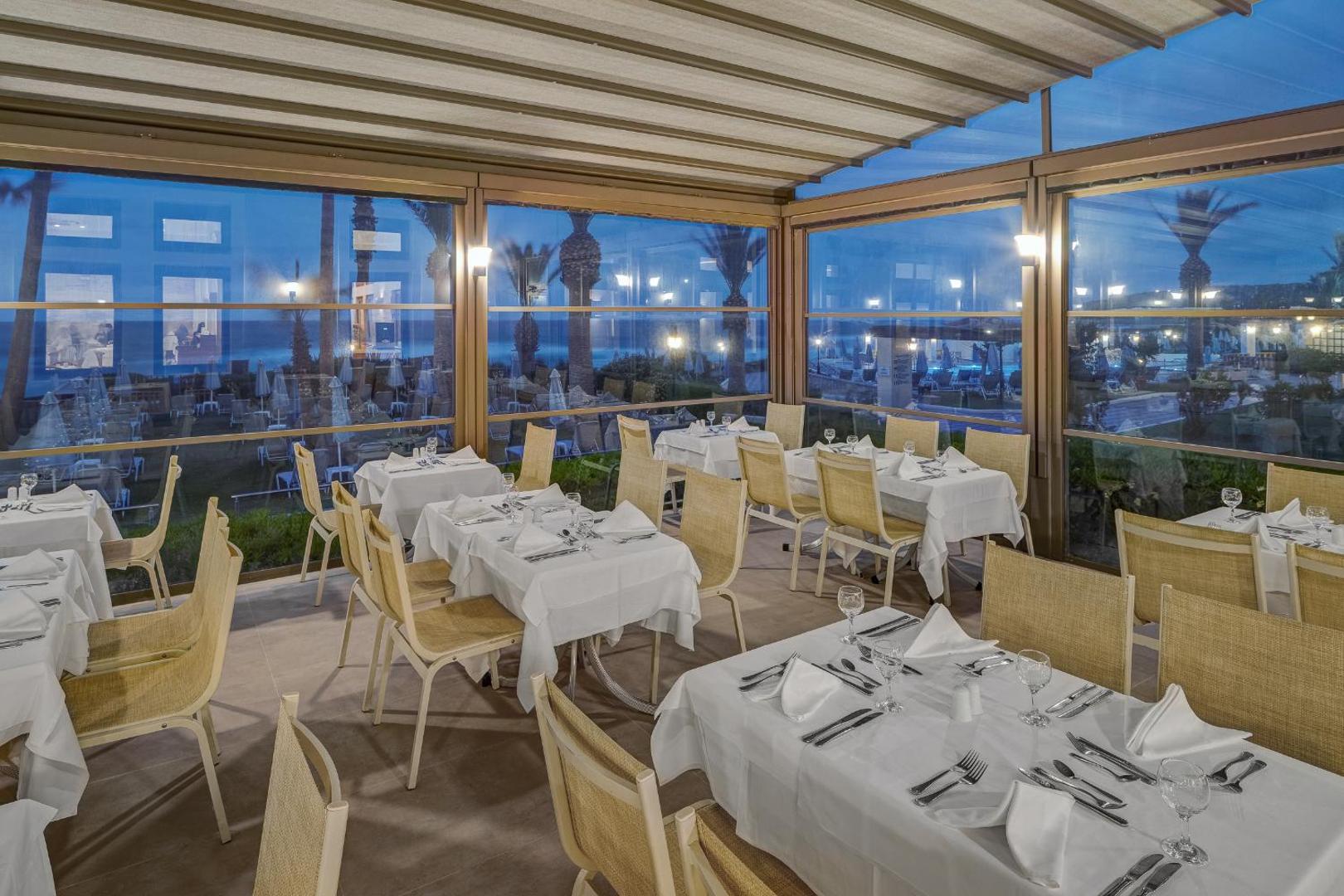 Creta Star Hotel iestiklota restorāna zāle vakarā ar klātiem galdiem un skatu uz izgaismoto viesnīcas teritoriju un jūru.