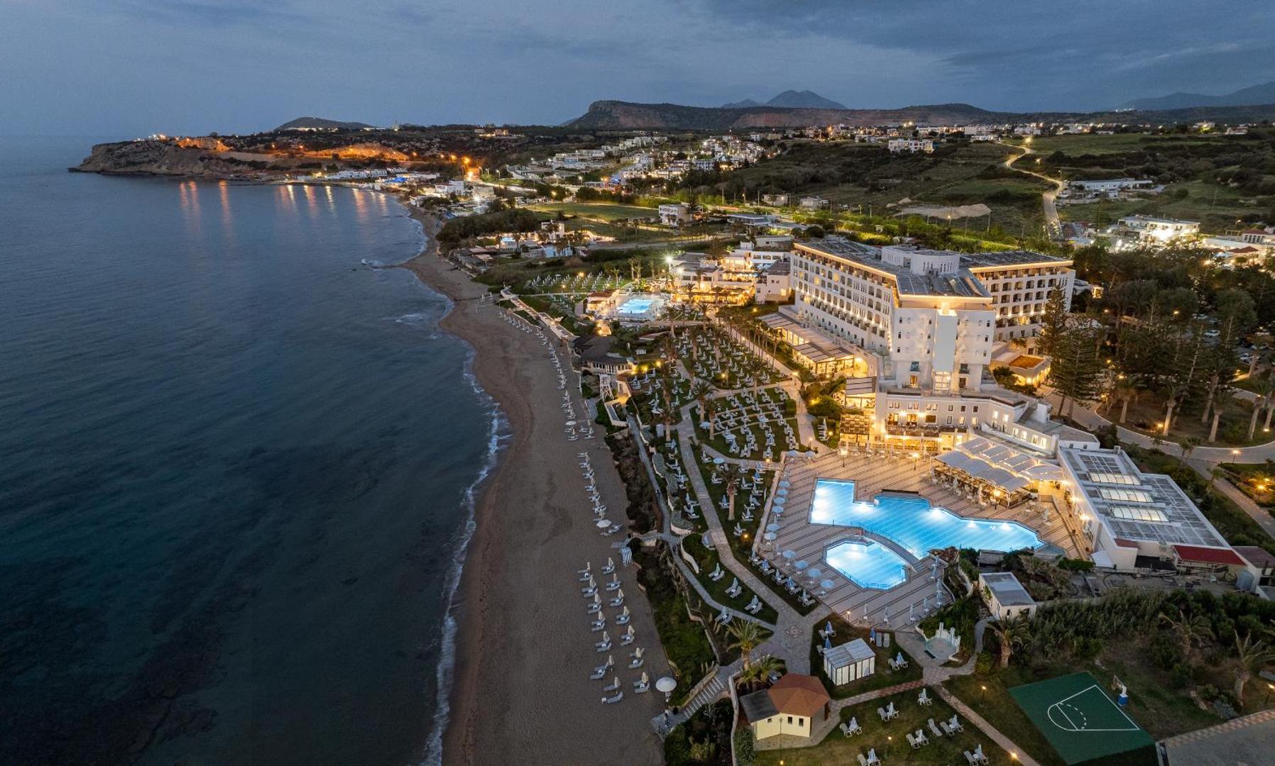 Creta Star Hotel skats no putna lidojuma vakarā — izgaismota viesnīcas ēka, baseini un plaša smilšu pludmale Krētas piekrastē