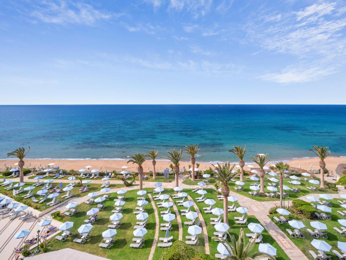 Creta Star Hotel dārzs ar daudzām palmām, saulessargiem un zālienu tieši pie smilšu pludmales un zilās Vidusjūras.
