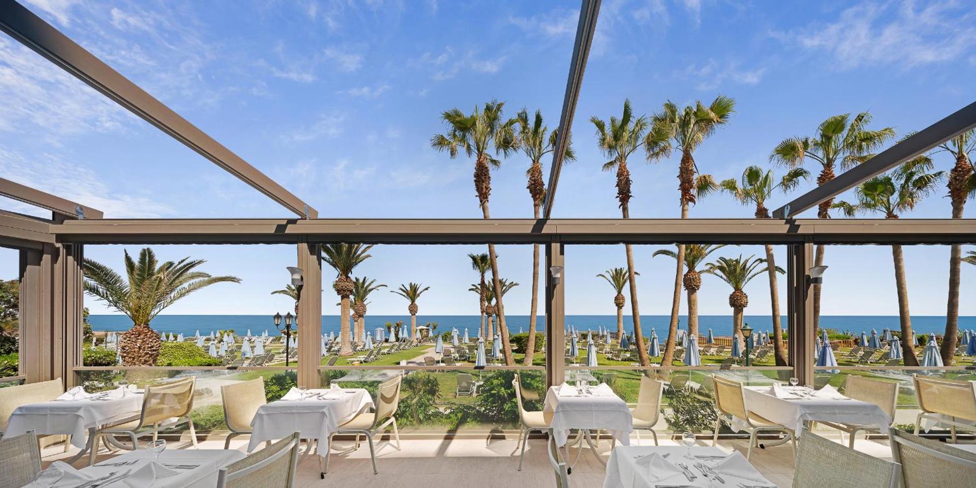 Creta Star Hotel restorāna terase ar klātiem galdiem, skatu uz palmām un jūru caur lieliem panorāmas logiem dienas laikā.