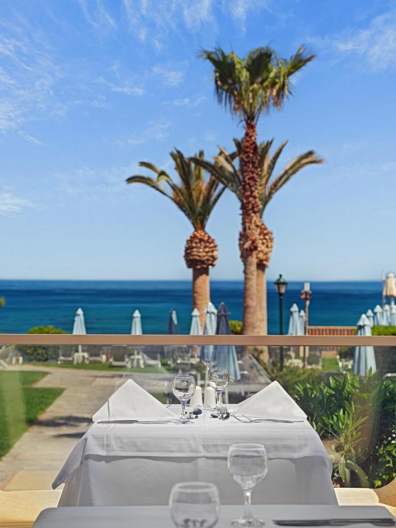 Creta Star Hotel restorāna galdiņš divām personām uz terases ar skatu uz palmām un Vidusjūru saulainā dienā.