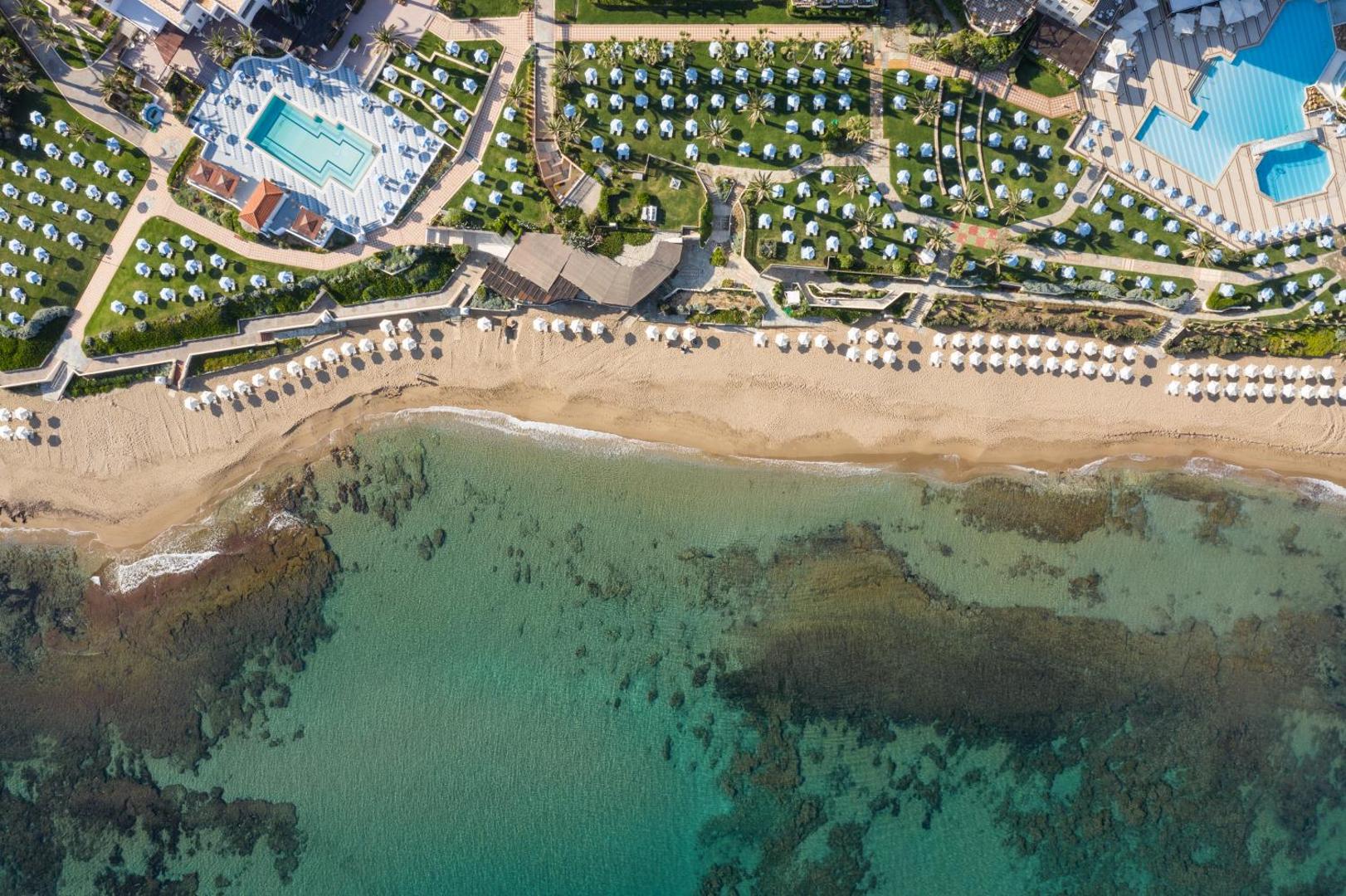 Skats no augšas uz Creta Star Hotel pludmali un zālienu ar saulessargiem — kristāldzidrs ūdenis un sakopta atpūtas zona.