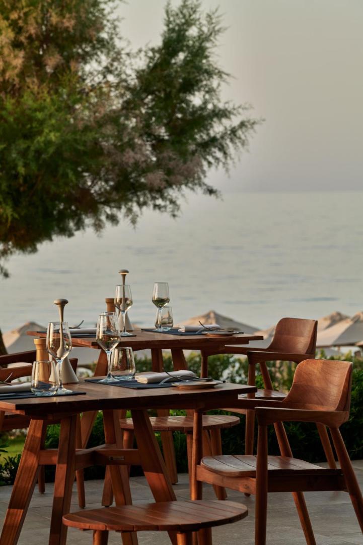 Amira Luxury Resort restorāna āra terase ar koka galdiem un krēsliem piekrastē, skats uz jūru saulrieta laikā