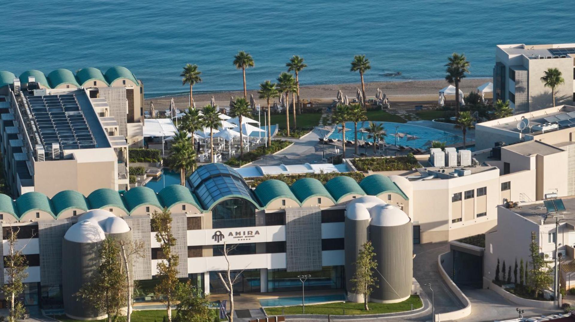 Amira Luxury Resort modernā arhitektūra un fasāde dienas gaismā — skats uz jūru, palmām un āra baseinu zonu