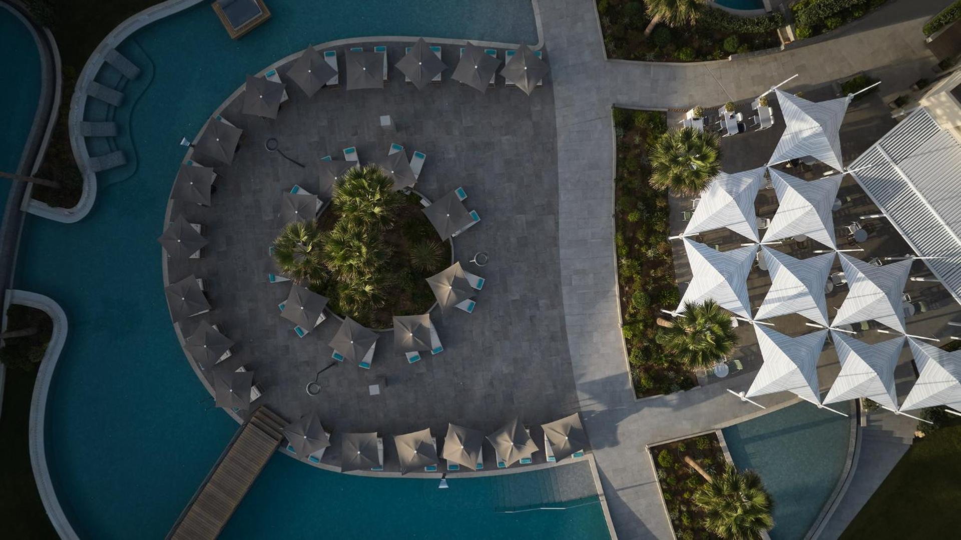 Amira Luxury Resort skats no augšas uz apaļo atpūtas zonu ar saulessargiem un palmām pie baseina Adelianos Kampos, Krētā