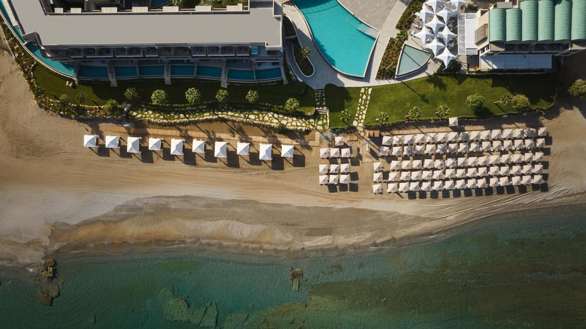 Amira Luxury Resort panorāmas skats uz privāto smilšu pludmali ar saulessargiem un viesnīcas ēku pie tirkīzzilas jūras Krētā