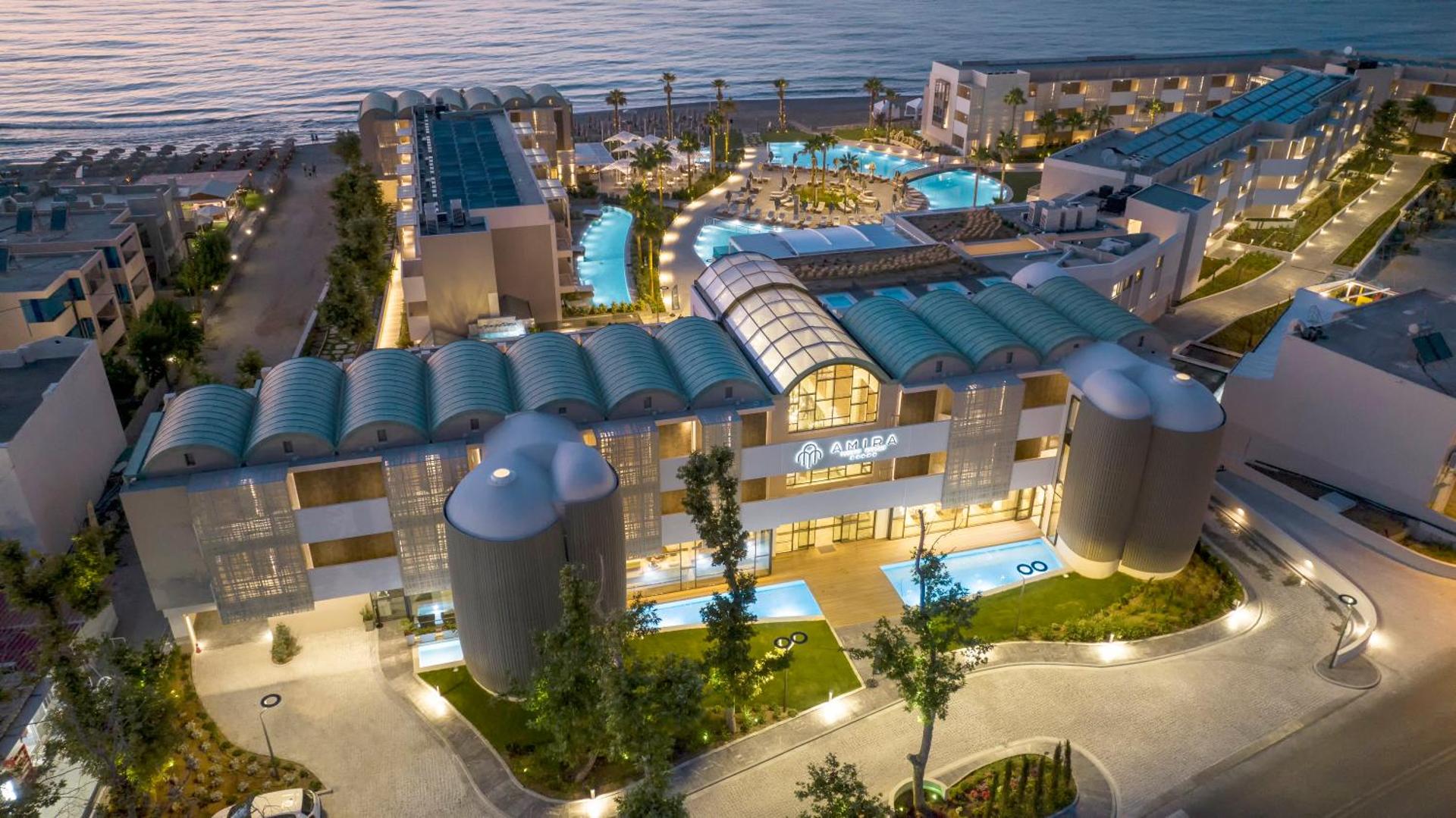 Amira Luxury Resort skats no putna lidojuma vakarā — izgaismota viesnīcas fasāde, baseini un piekrastes zona Retimno reģionā, Krētā