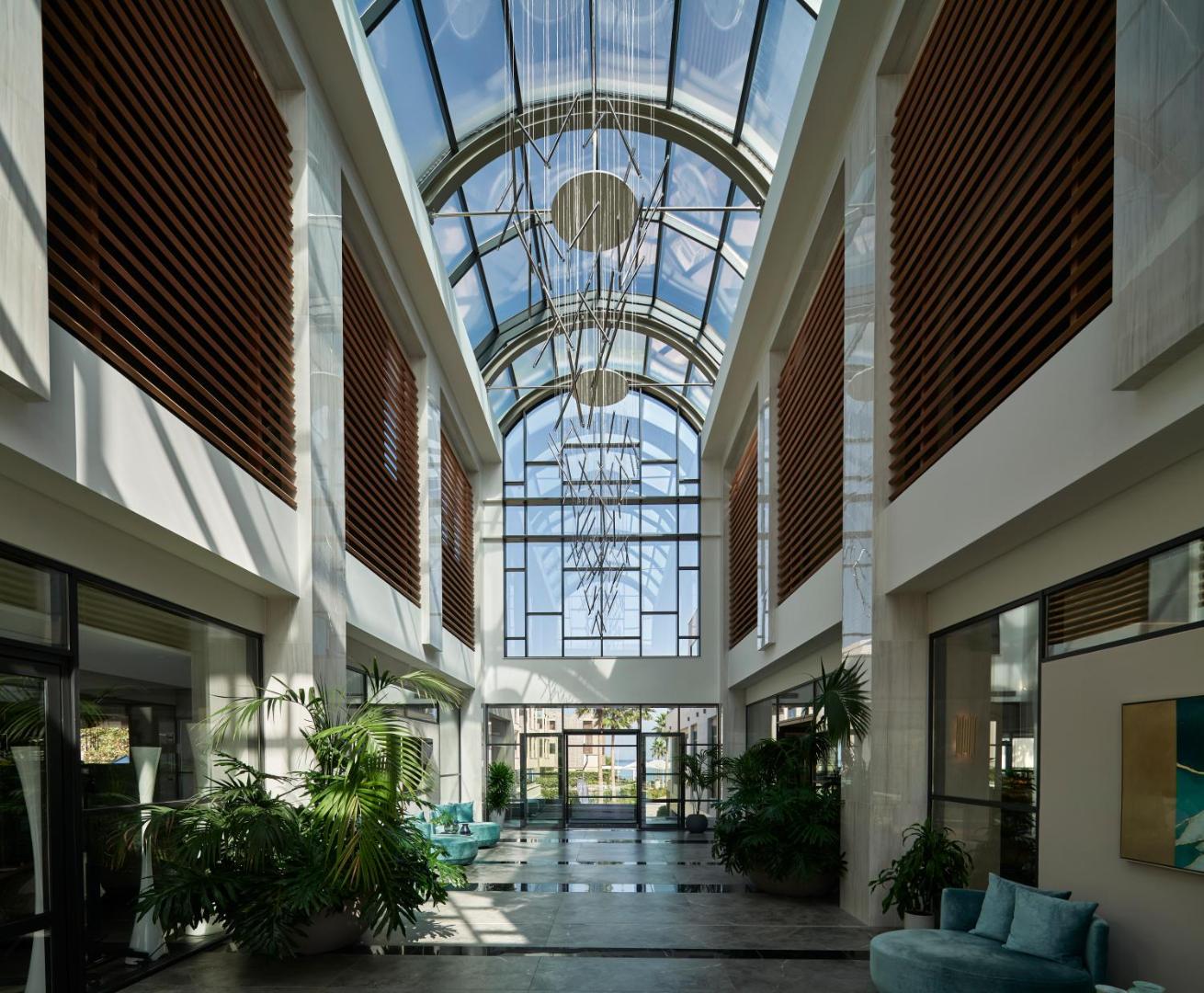 Amira Luxury Resort viesnīcas lobby ar augstiem stikla griestiem, modernu lustru un telpaugiem, skats uz ieeju