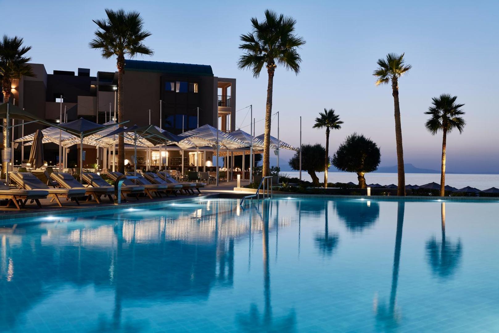 Amira Luxury Resort āra baseins vakarā ar izgaismotiem sauļošanās krēsliem, palmām un restorāna zonu piekrastē