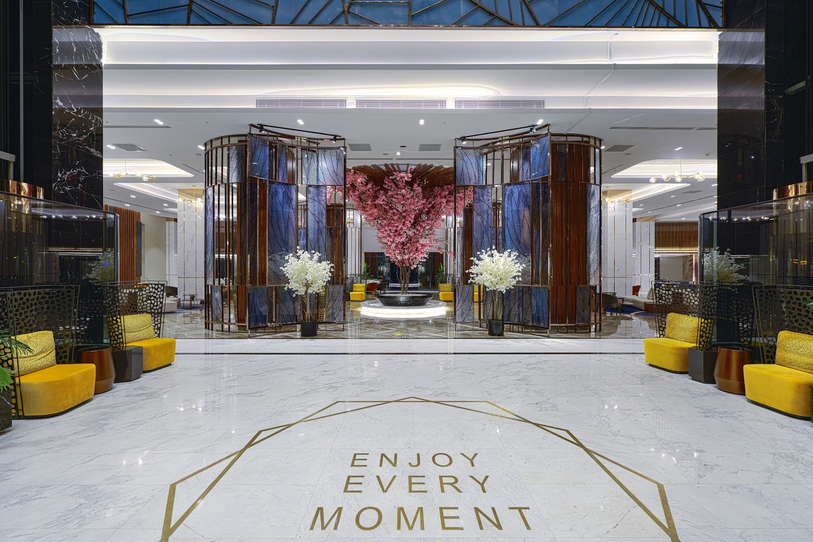 Kirman Calyptus Resort & SPA recepcijas zona ar dekoratīviem ziedu kokiem un uzrakstu Enjoy Every Moment uz marmora grīdas.