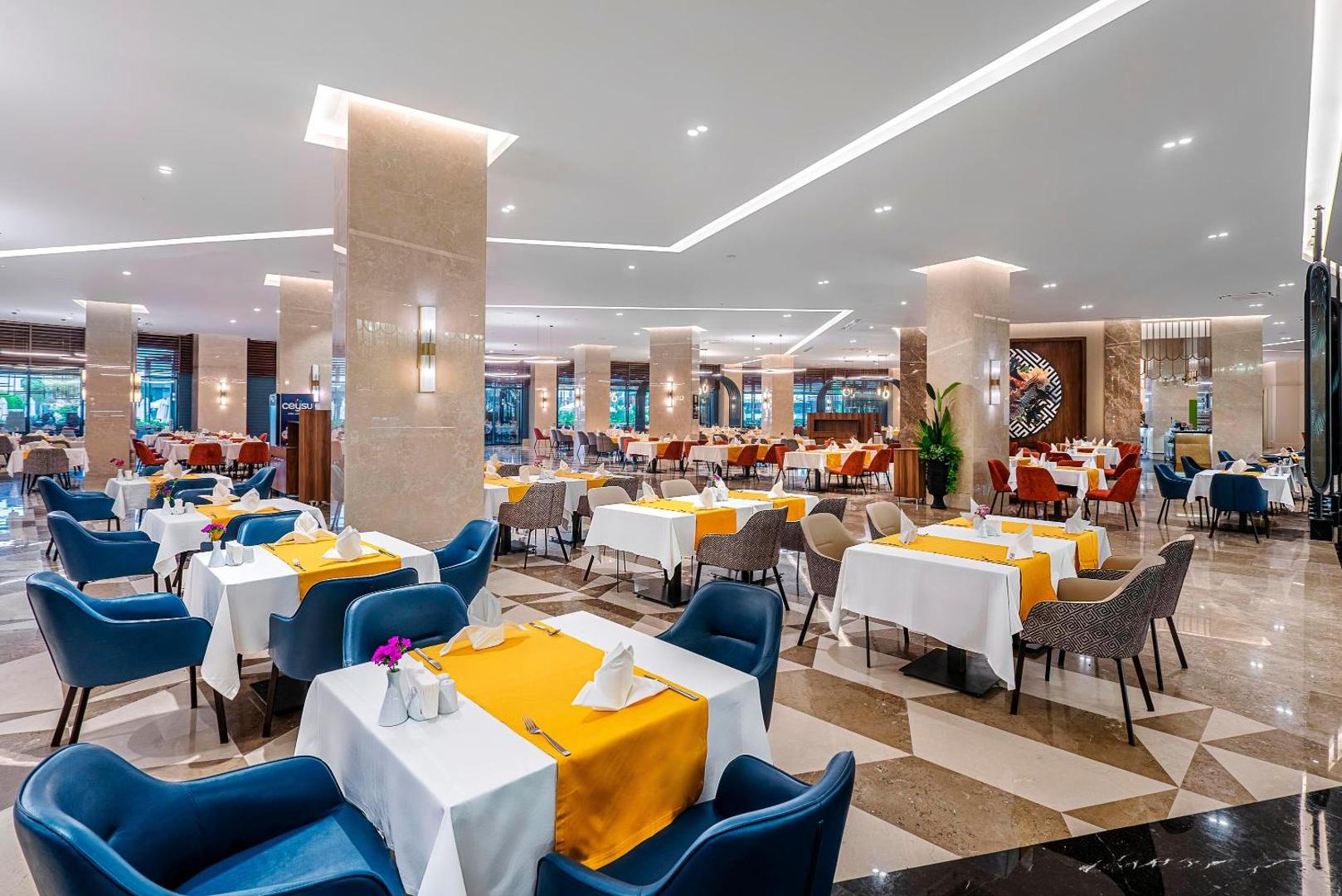 Kirman Calyptus Resort & SPA galvenais restorāns ar gaišu interjeru, marmora kolonnām un krāsainiem krēsliem