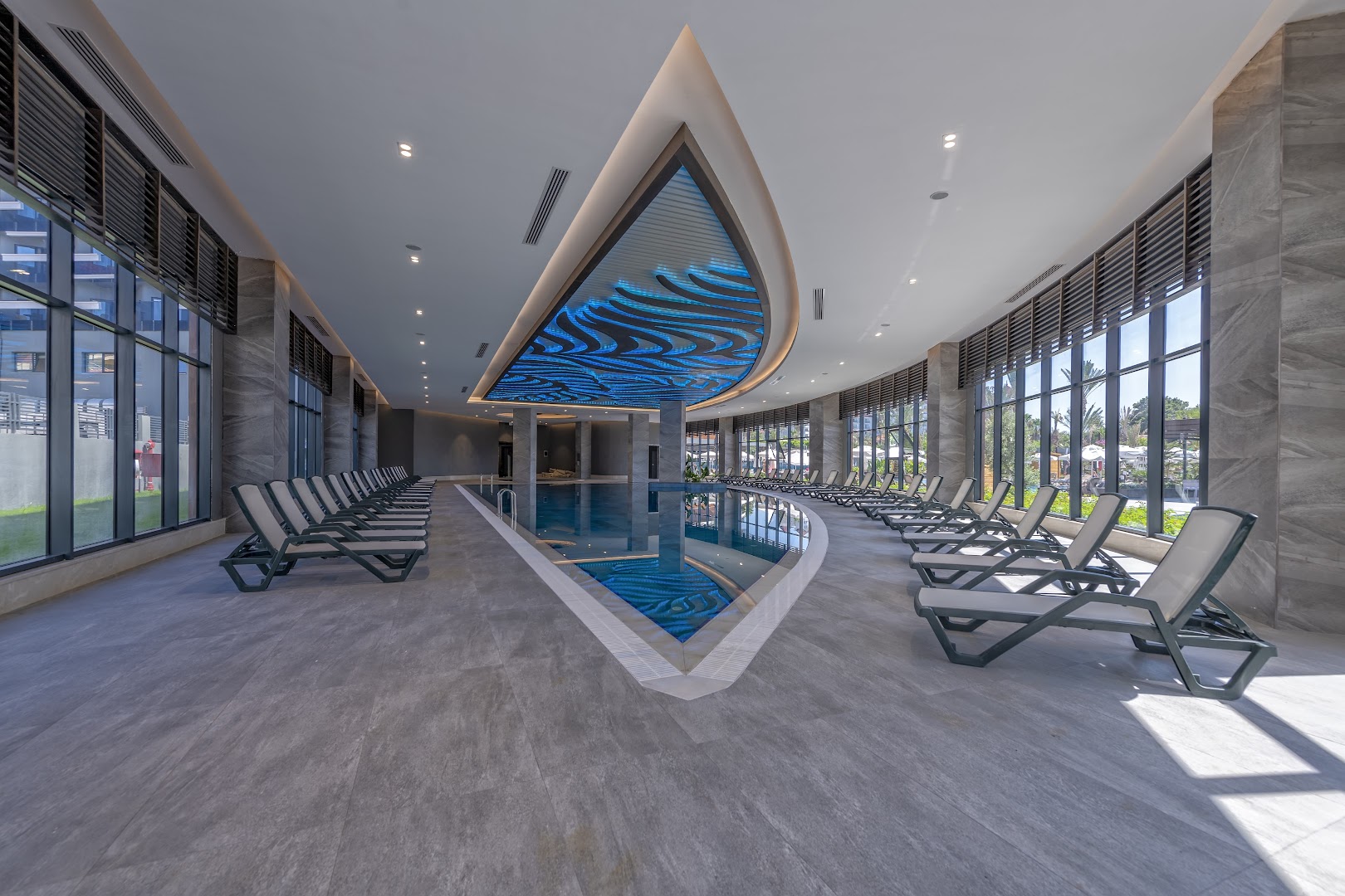 Plašs skats uz Kirman Calyptus Resort & SPA iekštelpu baseinu ar panorāmas logiem un daudziem atpūtas krēsliem.