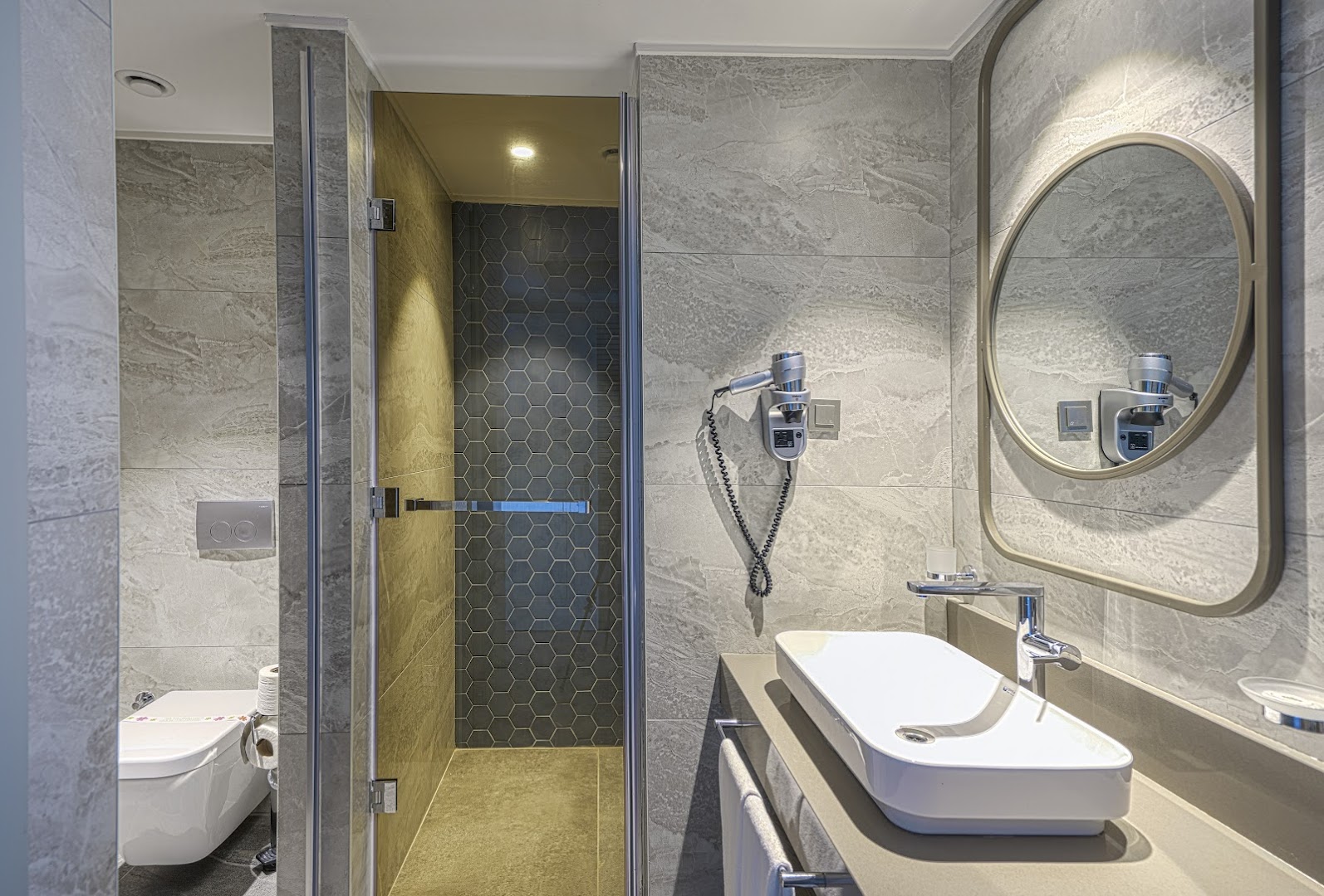 Kirman Calyptus Resort & SPA numura vannasistaba ar modernu dušas kabīni, akmens flīzēm, apaļu spoguli un fēnu