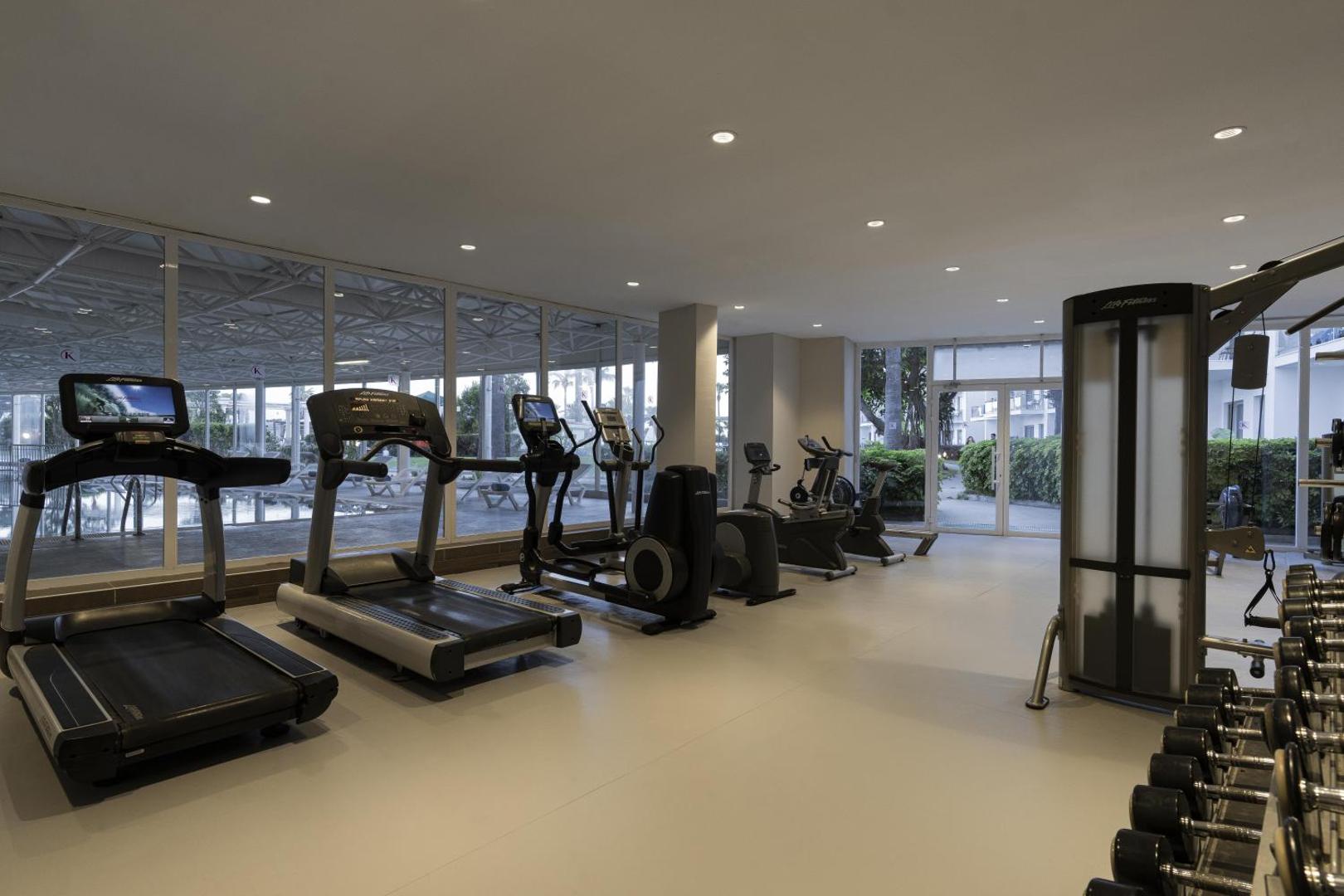 Kaya Side fitness centrs ar skrejceļiem un trenažieriem pie lieliem logiem ar skatu uz viesnīcas dārzu