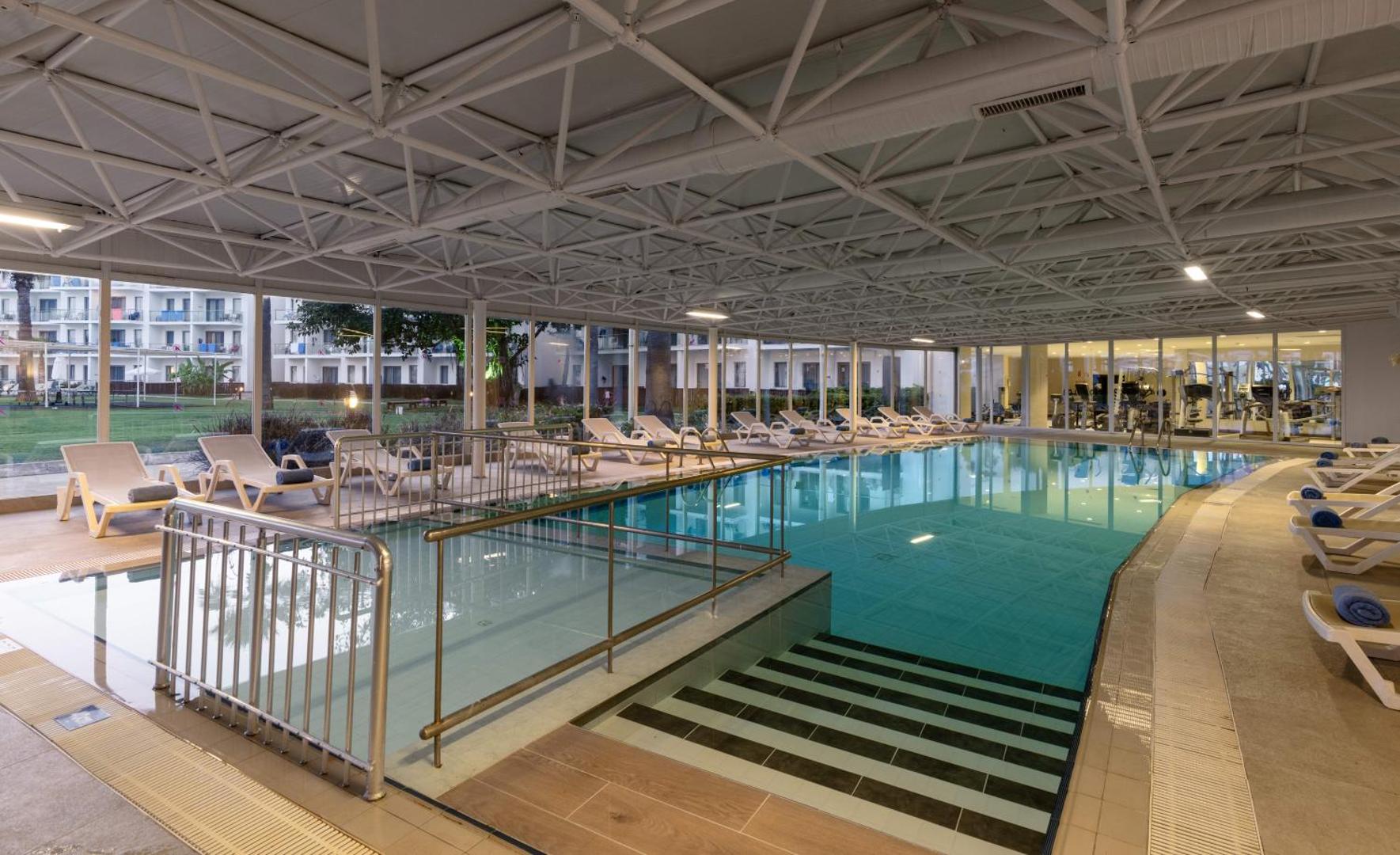 Kaya Side viesnīcas iekštelpu baseins ar panorāmas logiem un atpūtas krēsliem — gaiša spa zona ar skatu uz dārzu un fitnesa zāli fonā.