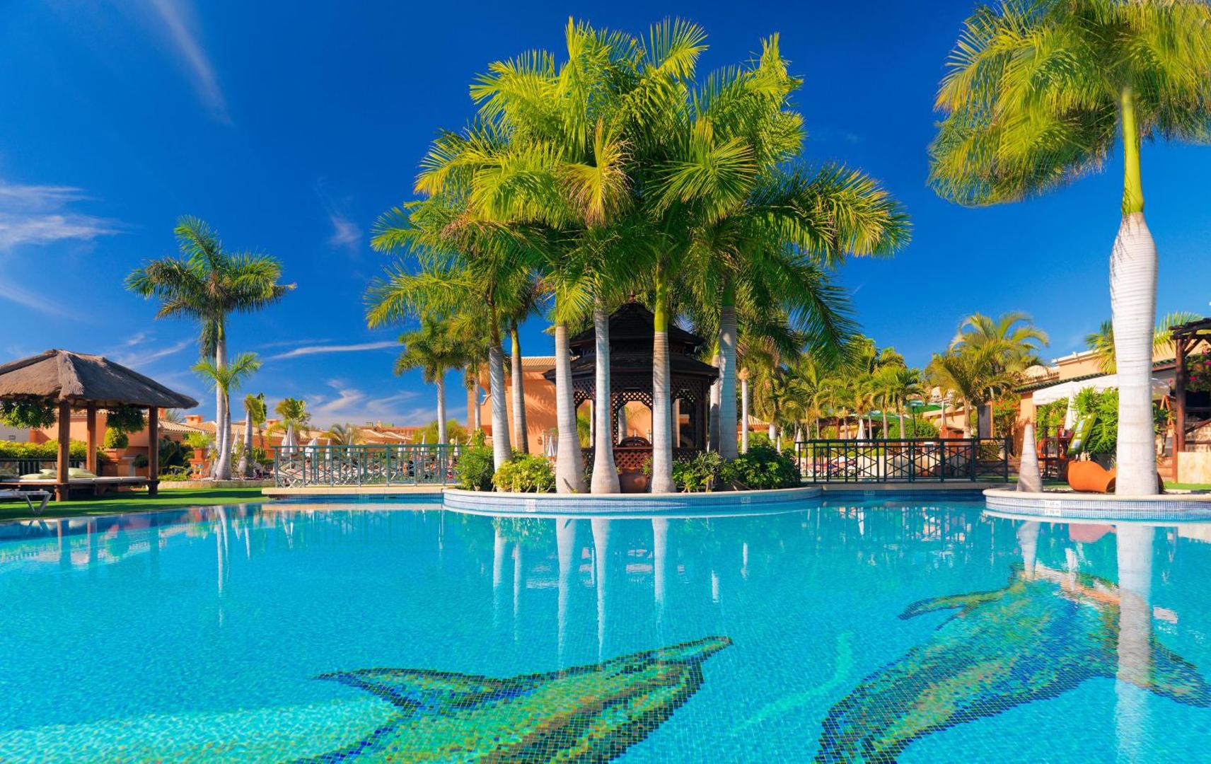 Green Garden Eco Resort galvenais baseins saulainā dienā ar palmām un koka lapeni — zils ūdens ar delfīnu zīmējumiem Tenerifes kūrortā