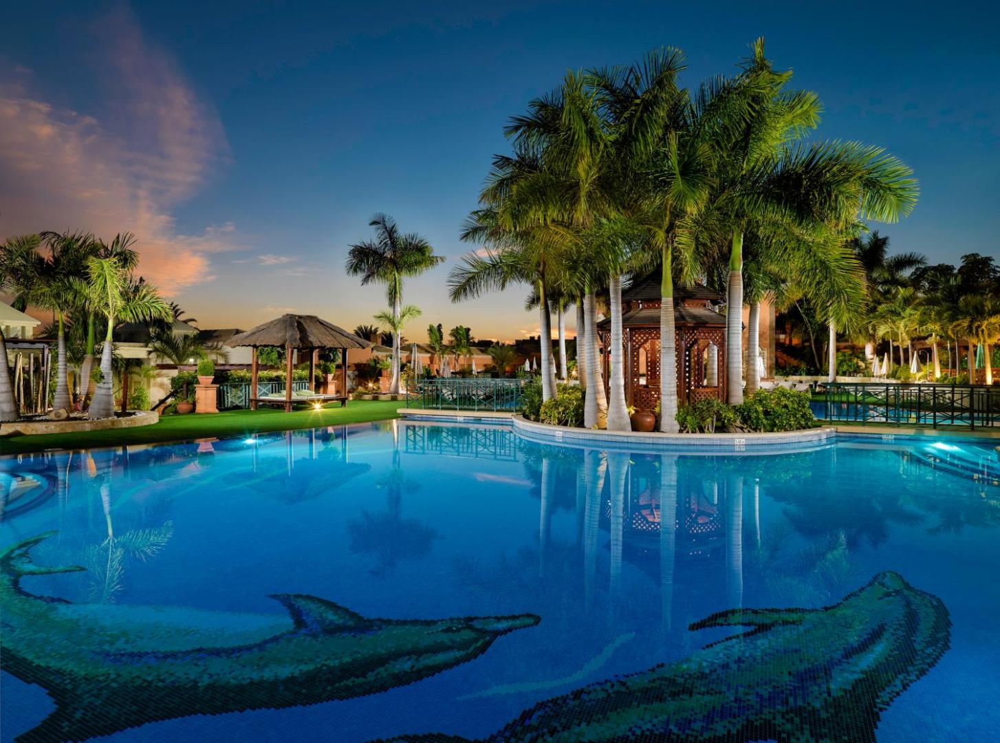 Green Garden Eco Resort āra baseins vakarā ar izgaismotām palmām un lapeni Tenerifē — mierpilna atmosfēra un delfīnu mozaīka ūdenī