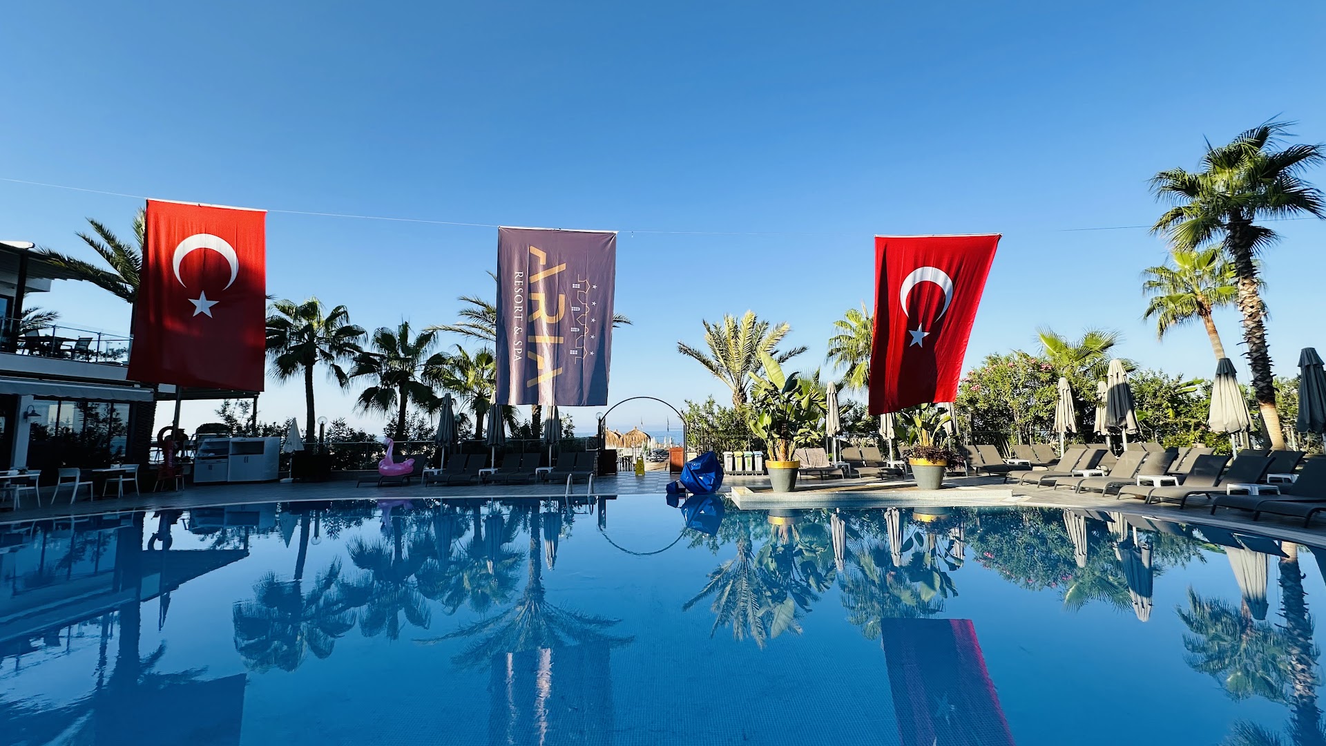 Aria Resort & Spa baseins ar Turcijas karogiem un viesnīcas simboliku, atspulgi ūdenī un sauļošanās krēsli
