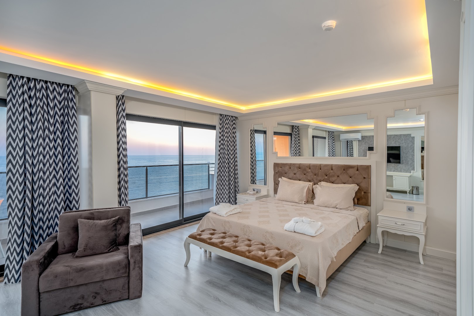 Aria Resort & Spa gaišs numurs ar divguļamo gultu, atpūtas krēslu un balkonu ar skatu uz jūru saulrietā Konakli reģionā