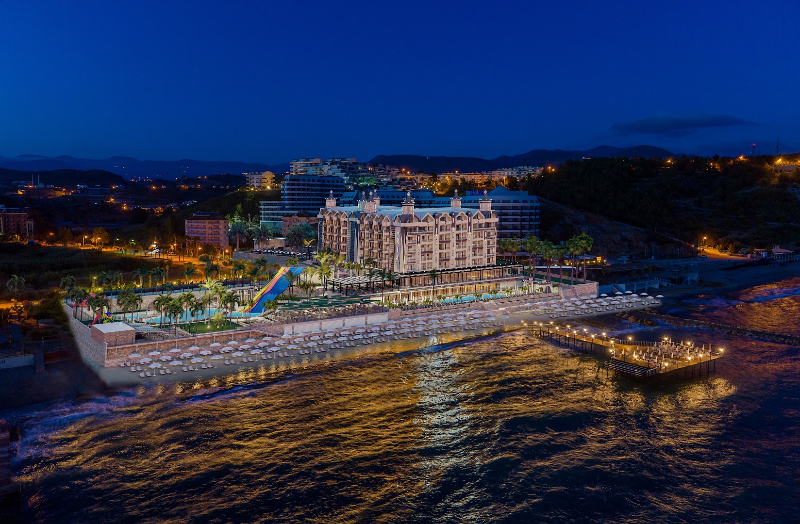 Aria Resort & Spa panorāmas skats naktī uz viesnīcas teritoriju, pludmali un izgaismoto piestātni Vidusjūras piekrastē