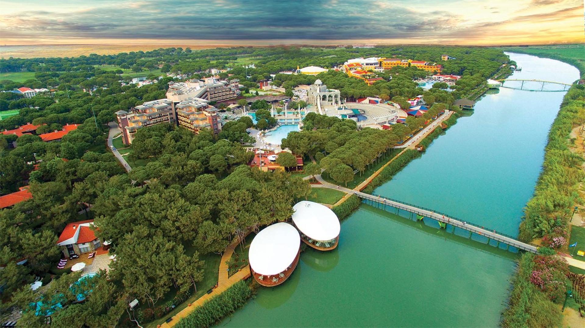 Xanadu Resort Hotel aerofotoattēls Belekā — plaša teritorija ar priežu mežu, upi, tiltu un baseinu kompleksu pie Antaljas piekrastes