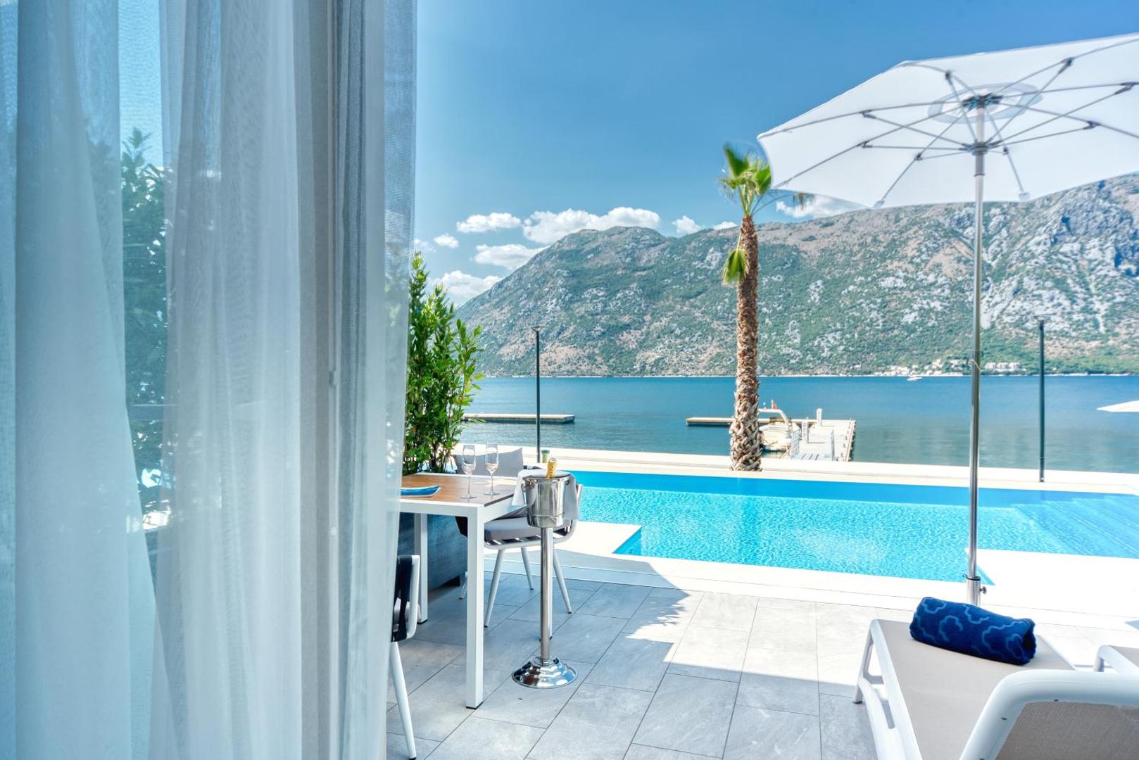 Skats no Hyatt Regency Kotor Bay Resort numura terases uz āra baseinu, palmu un Kotoras līča piekrasti saulainā dienā