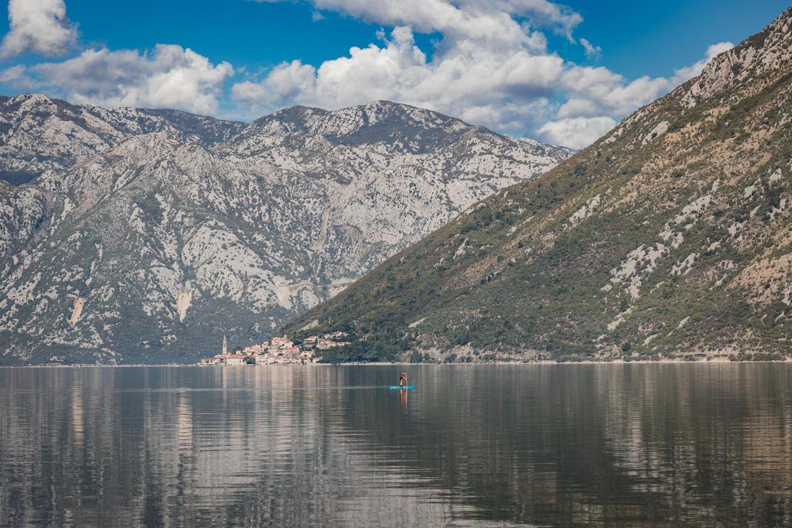 Hyatt Regency Kotor Bay Resort apkārtne ar mierīgu līča ūdeni, SUP dēļotāju un majestātiskiem kalniem tālumā