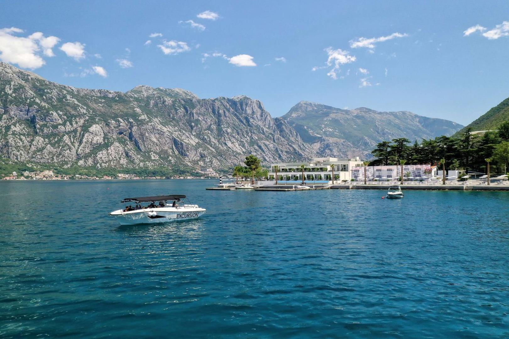 Skats no jūras uz Hyatt Regency Kotor Bay Resort piekrastē pie kalniem Melnkalnē — viesnīcas ēka un laiva līcī