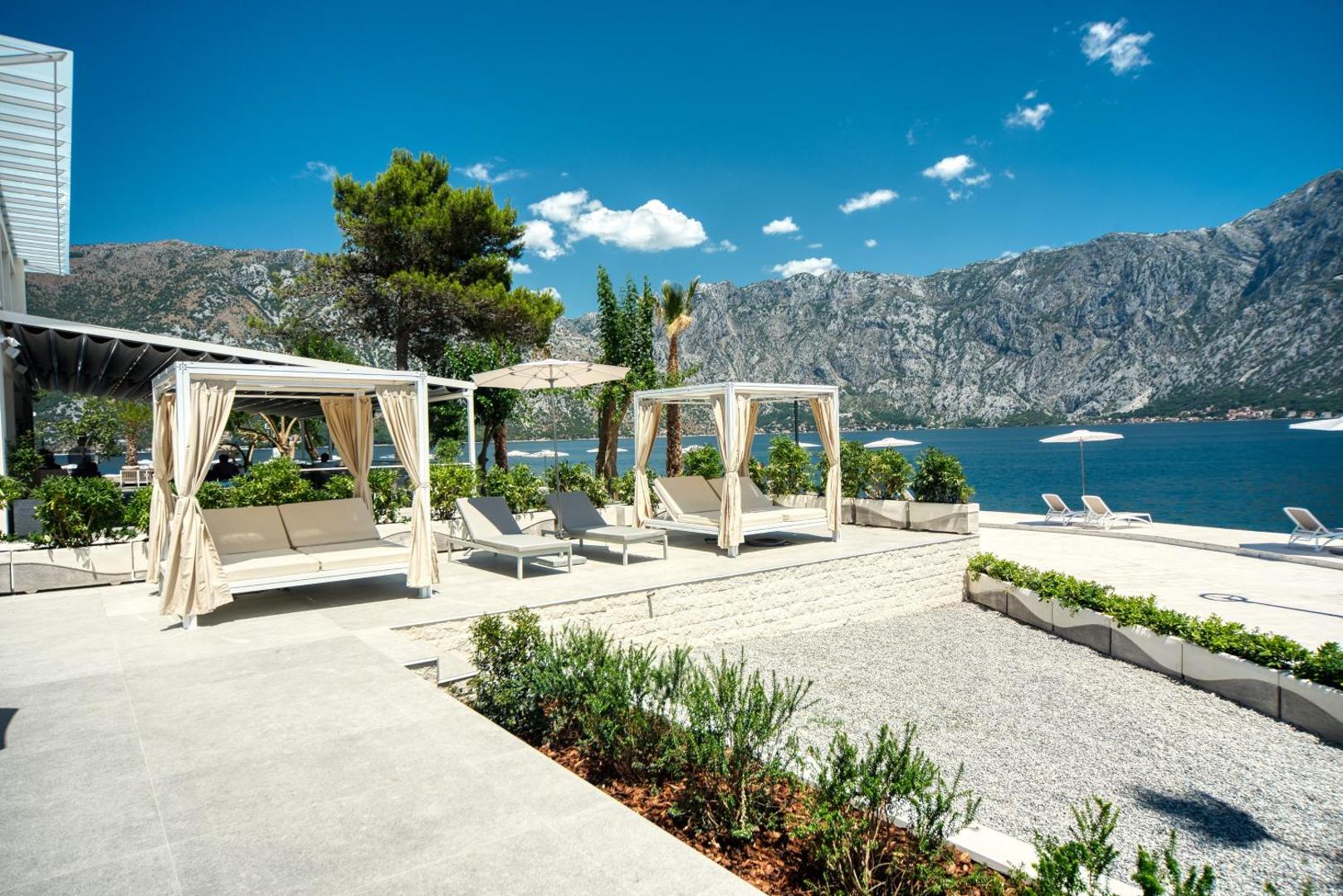 Hyatt Regency Kotor Bay Resort atpūtas zona ar grezniem saulessargiem un gultām pie līča Melnkalnē, fonā augsti kalni un zila jūra