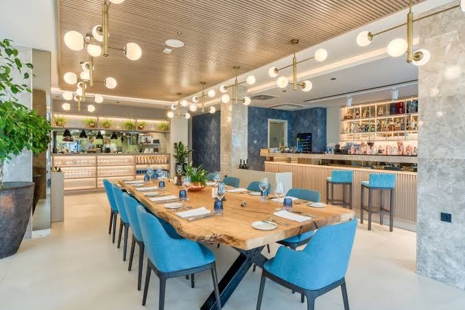 Hyatt Regency Kotor Bay Resort restorāns ar garu koka galdu, zilām mēbelēm un modernu apgaismojumu Melnkalnē — eleganta vakariņu zona