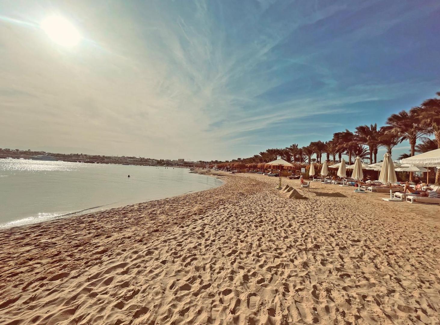 Sunrise Tucana Resort plašā pludmales zona ar gaišām smiltīm, saulessargiem un palmām pie dzidra ūdens Hurghada piekrastē