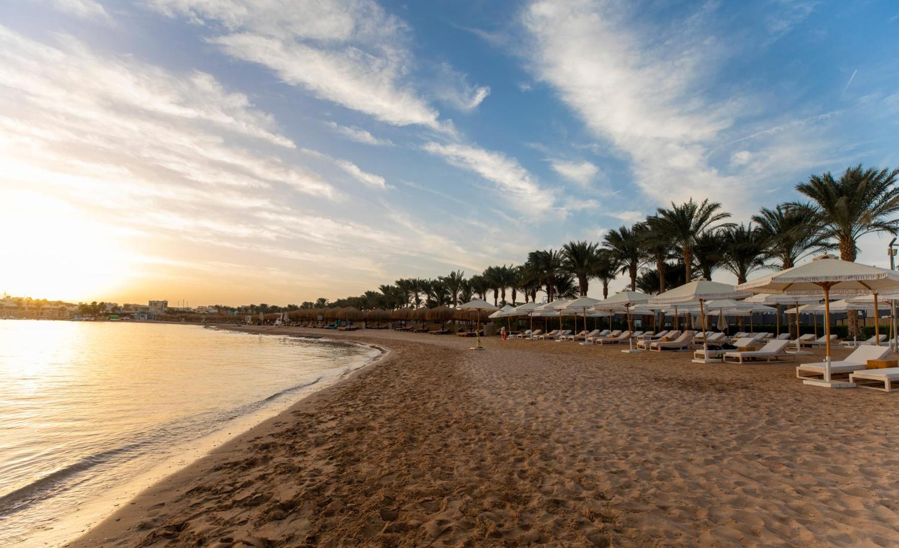 Sunrise Tucana Resort smilšu pludmale saulrietā Hurghada reģionā — rindas ar saulessargiem un atpūtas krēsliem pie mierīgas jūras un palmām