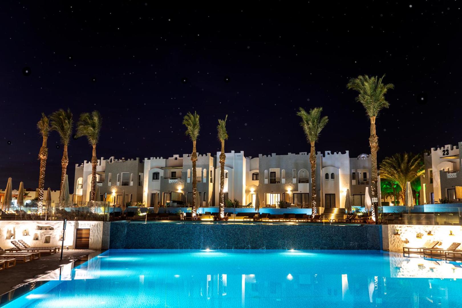 Sunrise Tucana Resort izgaismotais āra baseins un viesnīcas korpusi naktī, Hurgadā — zvaigžņotas debesis un palmu silueti pie ūdens.