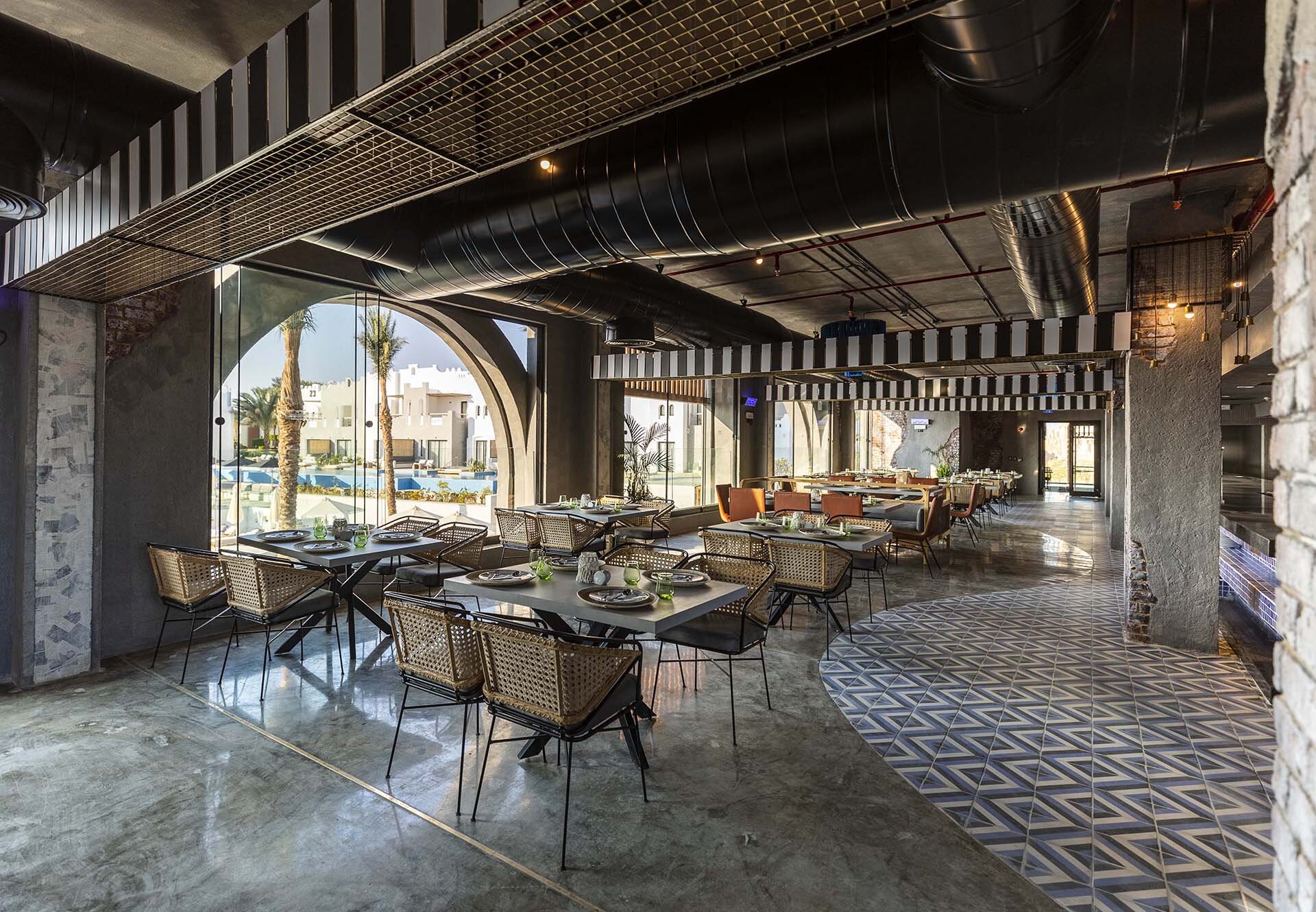 Sunrise Tucana Resort modernais restorāns ar industriāla stila dizainu, pītiem krēsliem un skatu caur lieliem logiem uz kūrorta teritoriju Hurgadā.