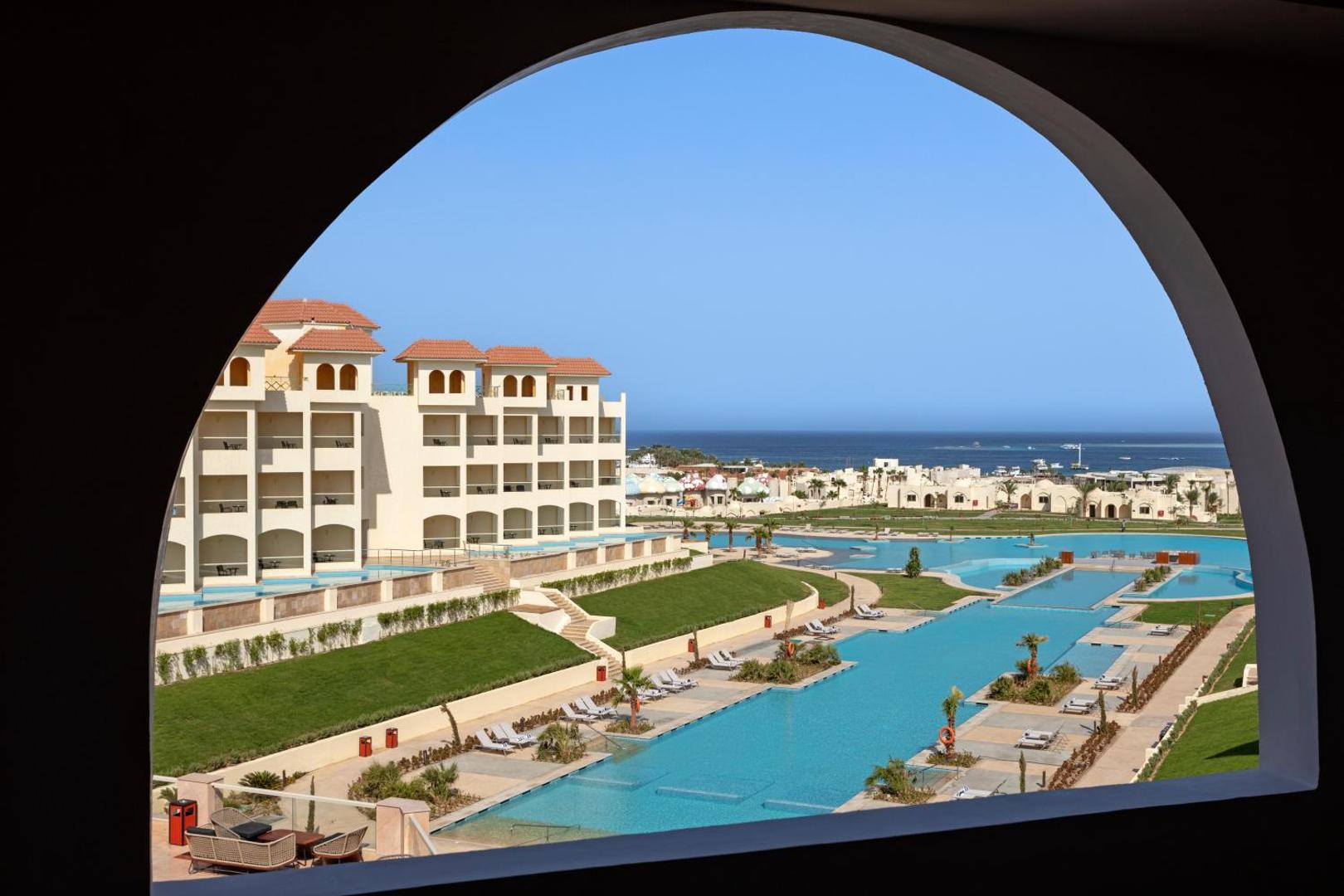 Skats caur arkas logu uz Xanadu Makadi Bay baseinu kompleksu, viesnīcas korpusiem un Sarkano jūru Makadi līcī