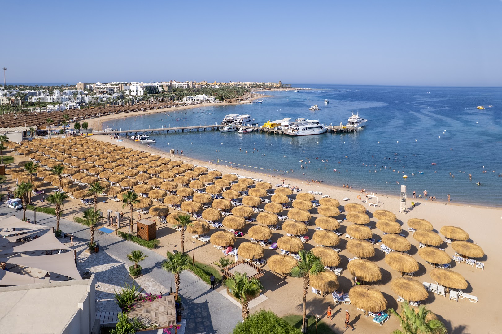 Xanadu Makadi Bay plašā smilšu pludmale ar salmu saulessargiem un piestātni Hurgadā — skats no augšas uz zilo līci