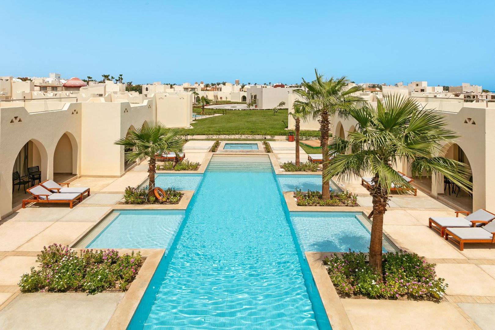 Xanadu Makadi Bay kūrorta baseins starp zemstāvu bungalo ēkām Hurgadā — gaiša arhitektūra, palmas un atpūtas krēsli pie ūdens