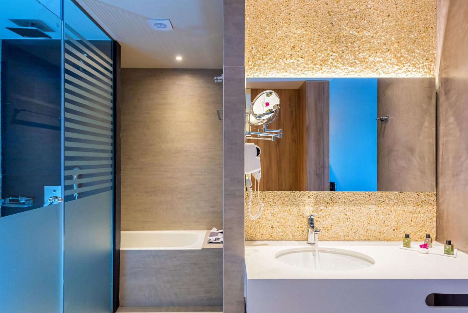 Solimar Aquamarine Resort numura vannasistaba ar modernu izlietni, lielu spoguli, vannu un dušas zonu Krētā — gaišs un mūsdienīgs dizains
