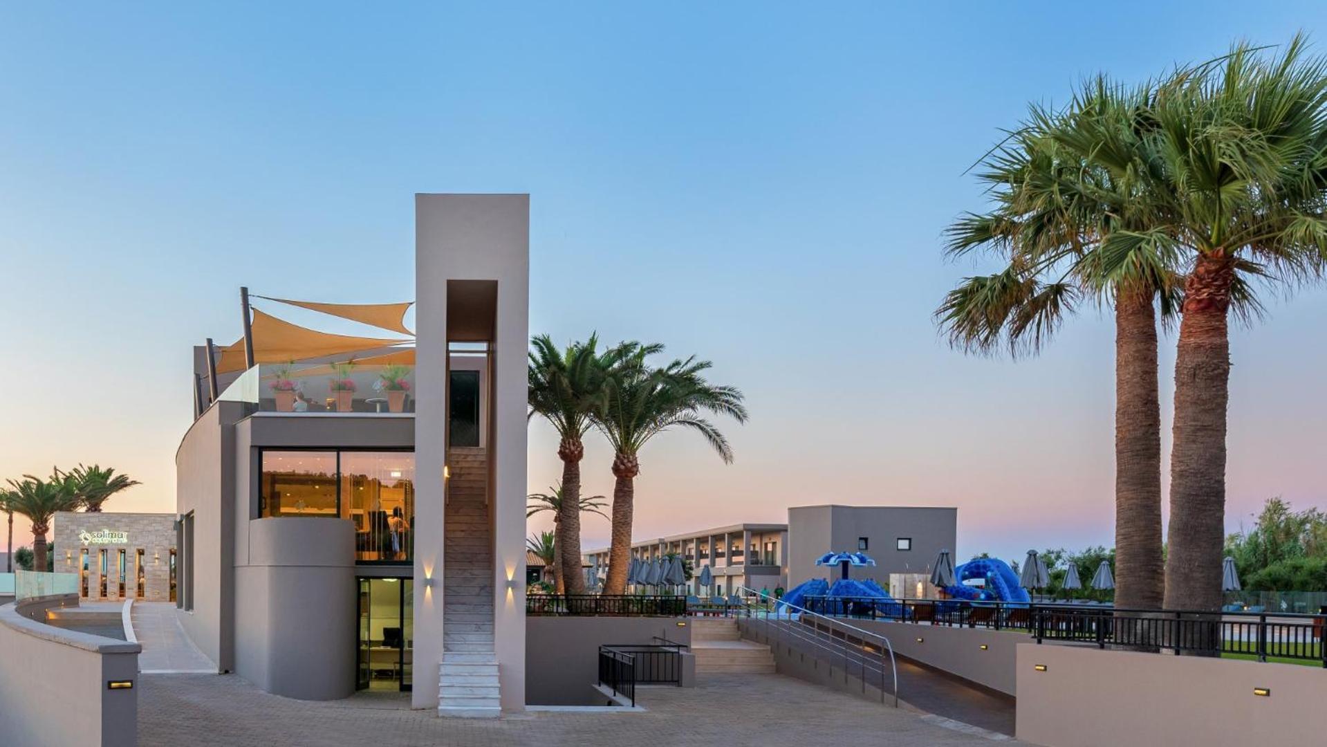 Solimar Aquamarine Resort modernā arhitektūra saulrietā ar palmām un bērnu ūdens atrakciju parku fonā Krētas salā