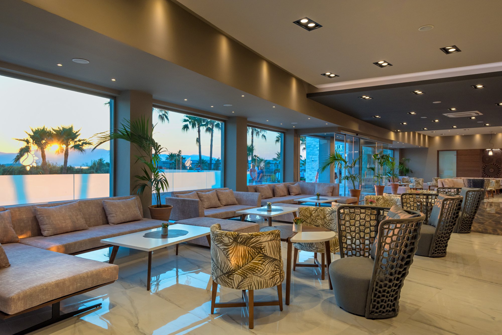 Solimar Aquamarine Resort modernais lobby bārs ar ērtiem dīvāniem, dizaina krēsliem un panorāmas logiem uz dārzu saulrieta gaismā