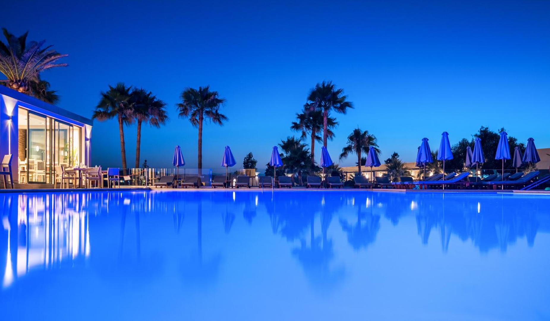 Solimar Aquamarine Resort āra baseins vakarā ar zilu apgaismojumu, palmām un saulessargiem piekrastē Krētā, Grieķijā