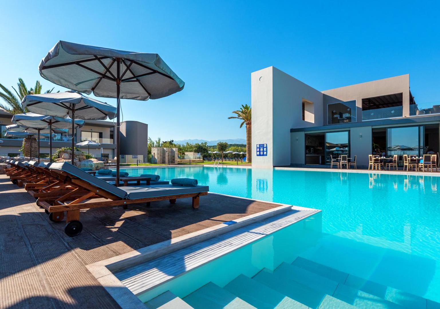 Solimar Aquamarine Resort lielais āra baseins ar sauļošanās krēsliem, saulessargiem un modernu arhitektūru saulainā dienā Krētā