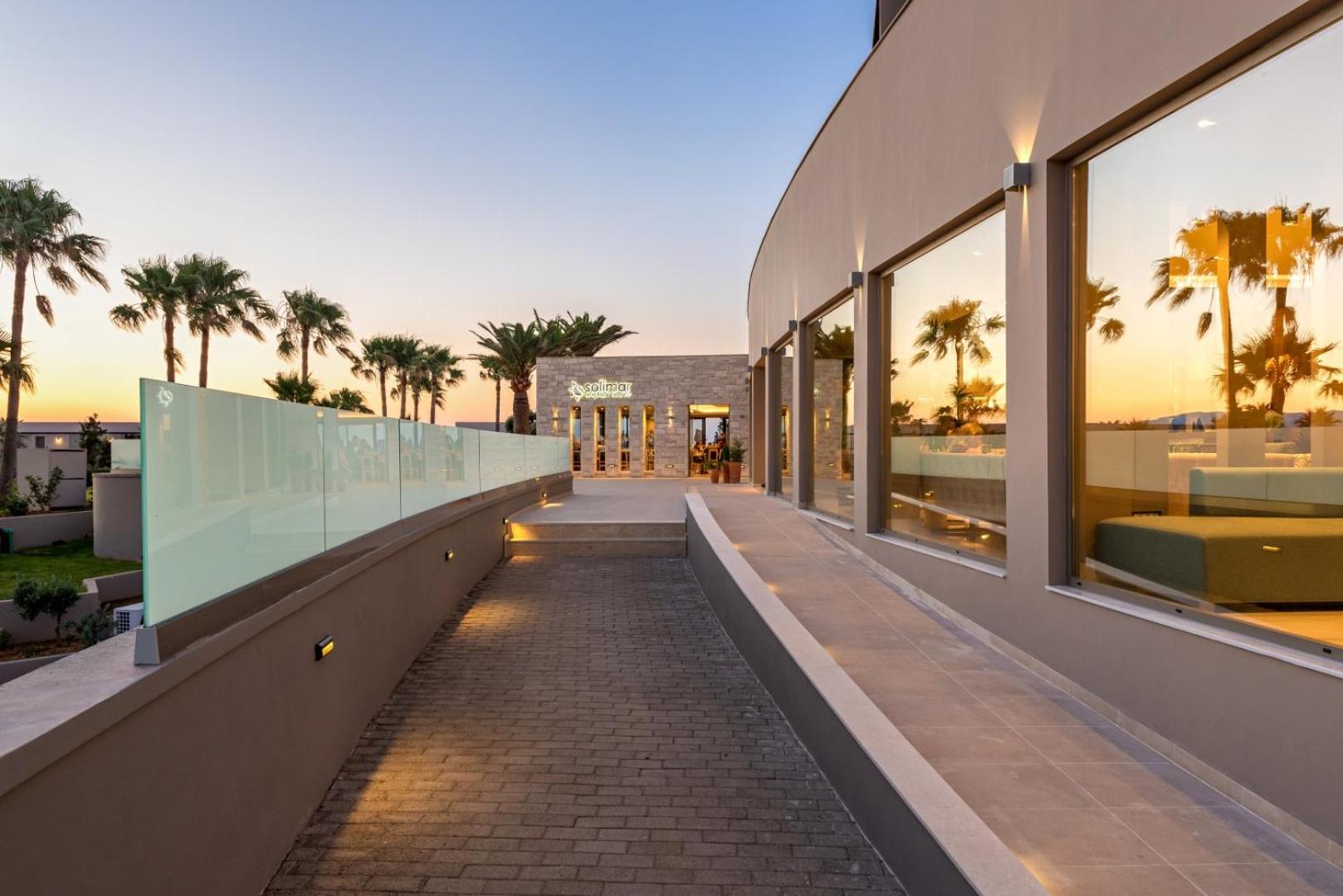 Solimar Aquamarine Resort restorāna ieeja un terase saulrieta gaismā ar stikla margām un palmām piekrastē