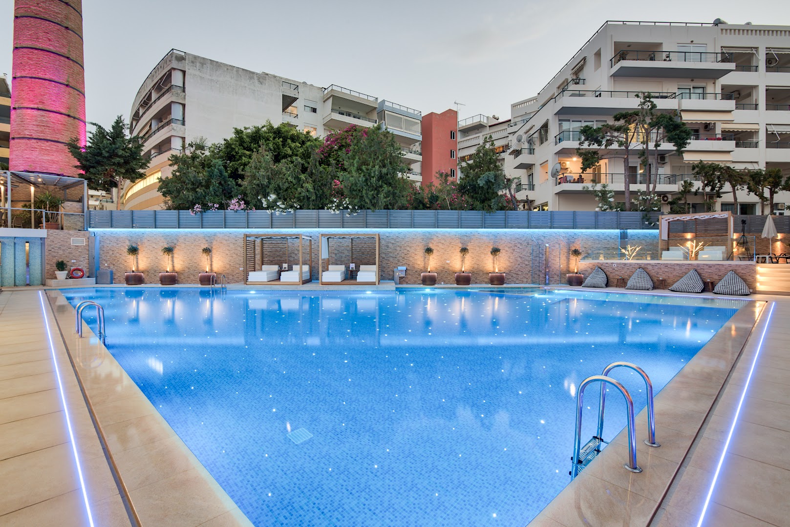 BIO Suites Hotel & Spa izgaismots baseins vakarā ar dekoratīvām gultām un pilsētas ēkām fonā — romantiska atmosfēra Retimno