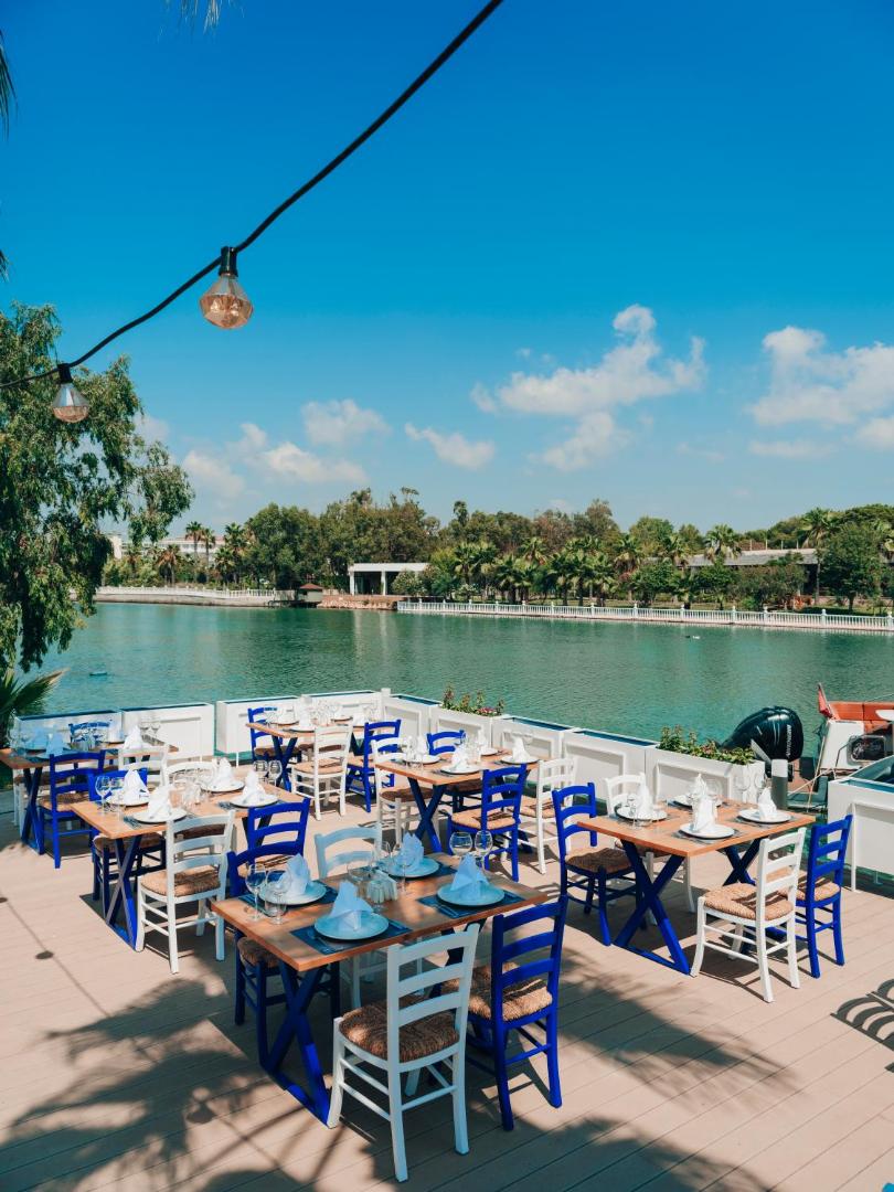 Selectum Family Resort Belek āra terases restorāns pie upes ar ziliem koka krēsliem un balti klātiem galdiem saulainā dienā